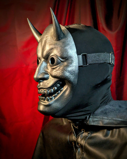 Side angle view of a person wearing a mask with horns and a dark cowl against a red curtain background