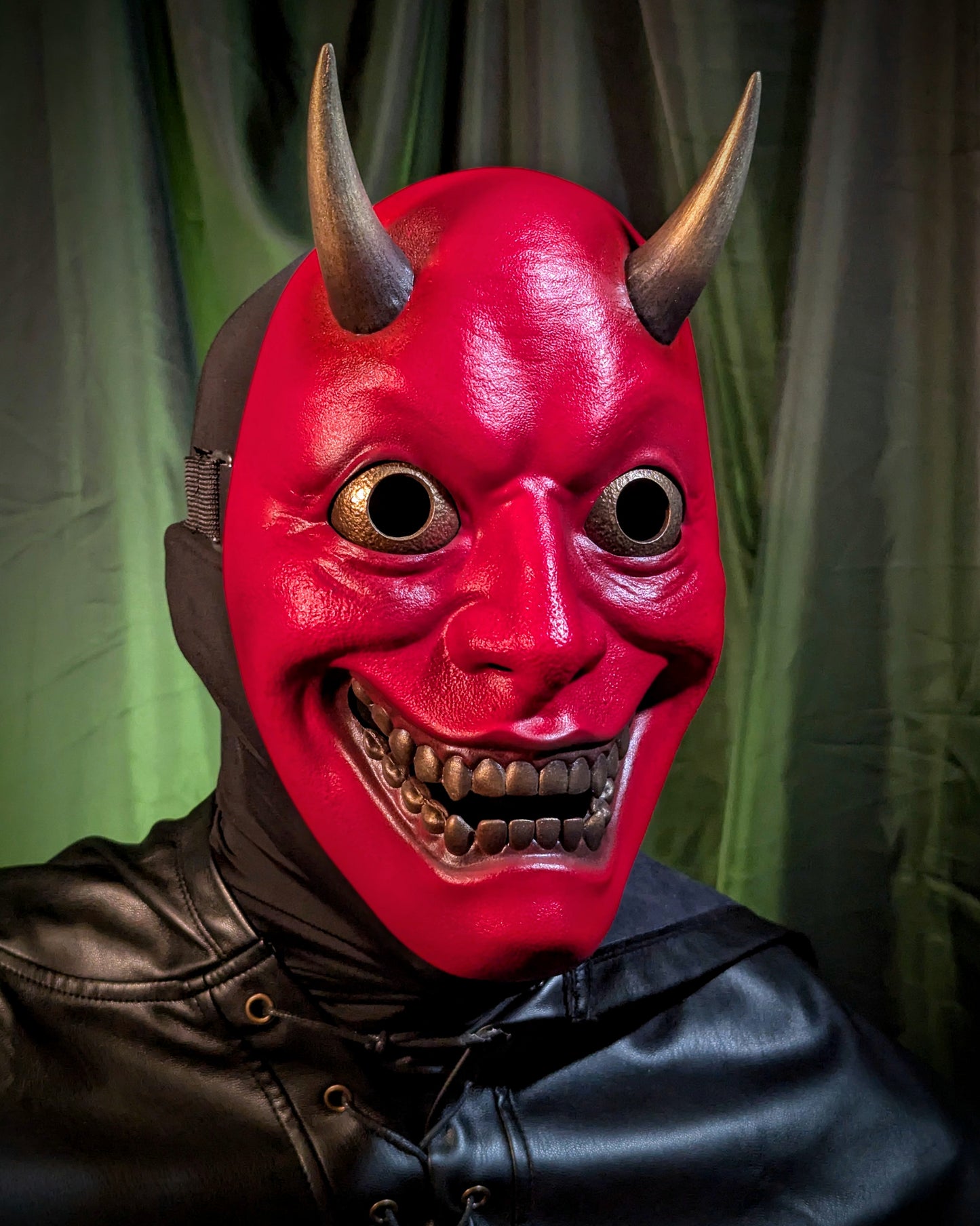 A closeup of a person wearing a red hannya mask with golden horns, eyes, and teeth against a green curtain background