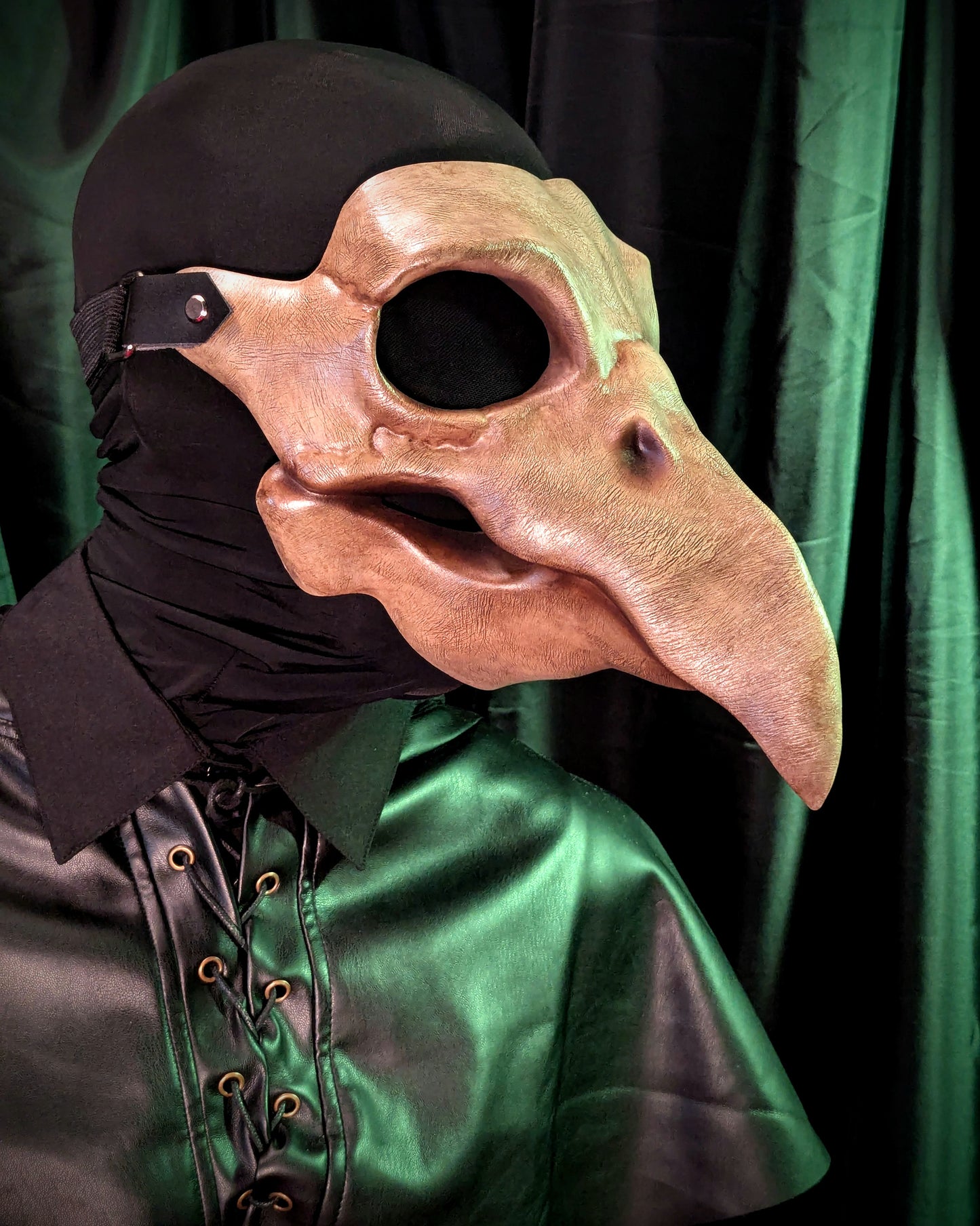 Person wearing a large bird-like mask with a long beak against a green curtain background