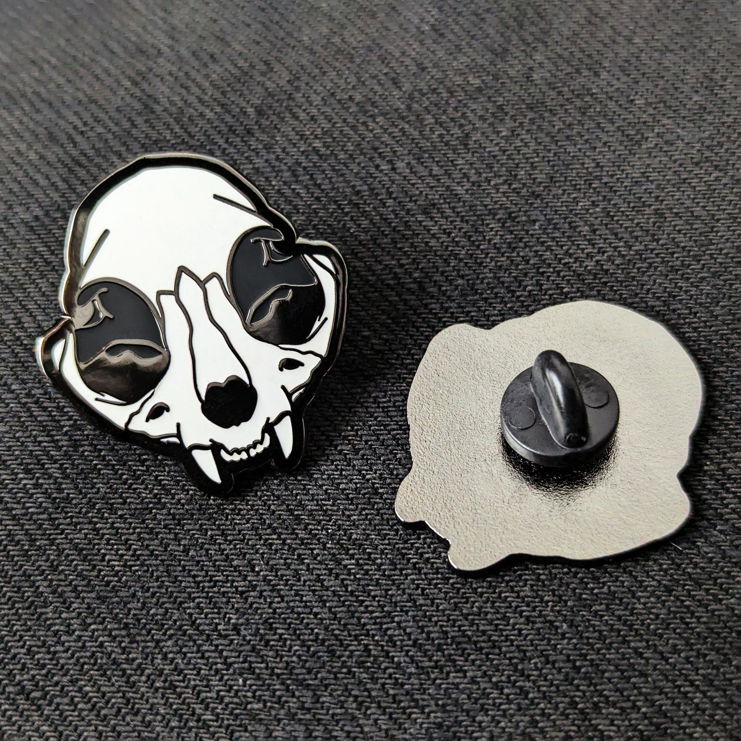 A hard enamel pin with a depiction of a cat skull with black nickel plating next to an identical pin facing downwards showing off a rubber backing