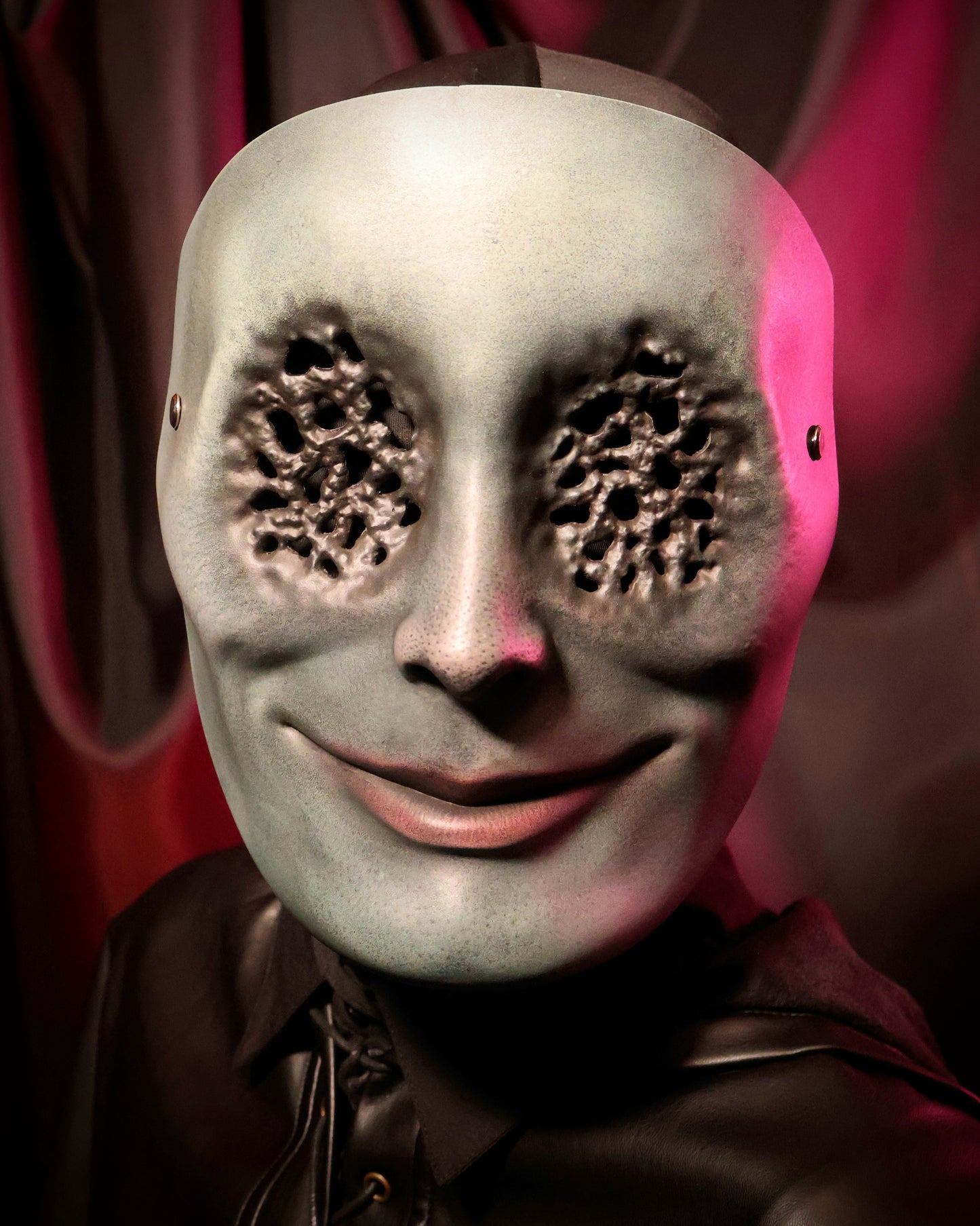 Front view of a person wearing a green mask with many small holes in the eyes on a dark background