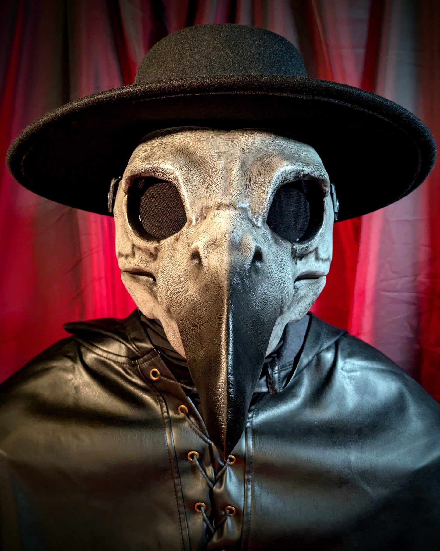 Person wearing a plague doctor mask and hat facing directly at the viewer against a red curtain background