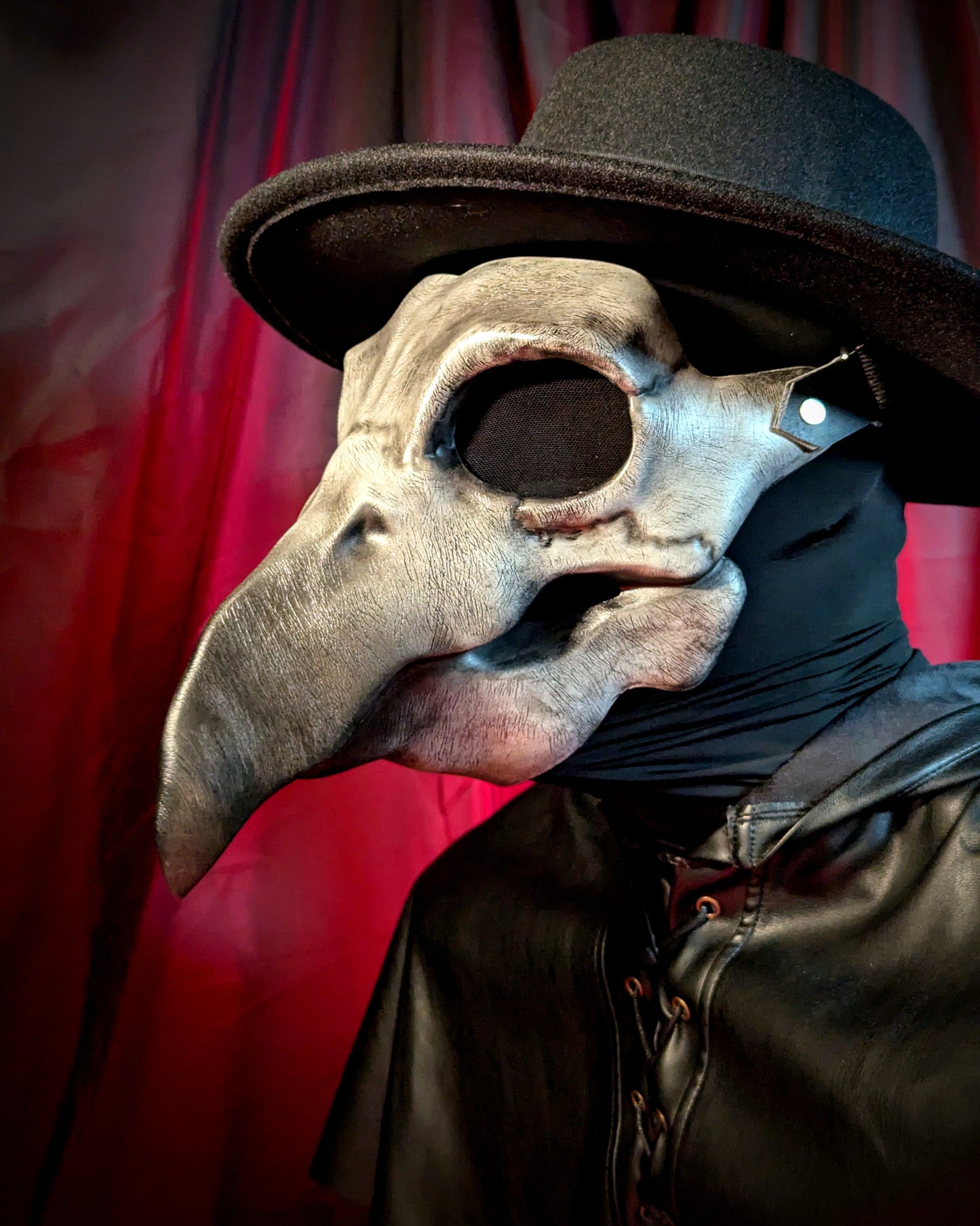 Person wearing a plague doctor mask with a long beak and black hat against a red curtain background