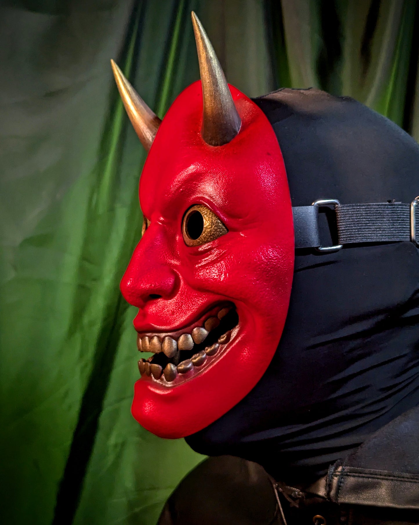 A side view of a person wearing a red hannya demon mask with horns against a green curtain background. This view shows off the side profile of the mask and the adjustable elastic head strap