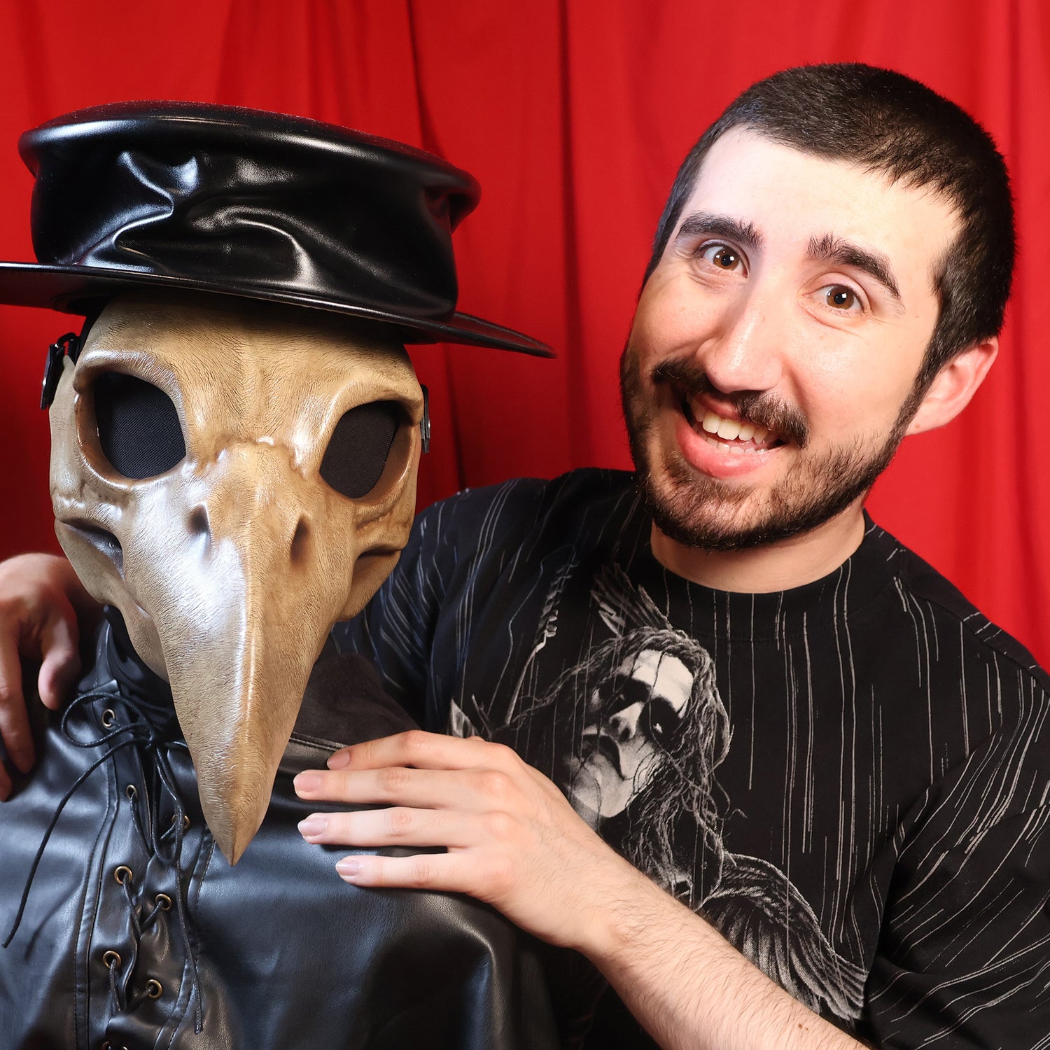 Deven Mendoza posing next to a bust wearing a black leather cowl, hat, and brown plague doctor mask against a red curtain