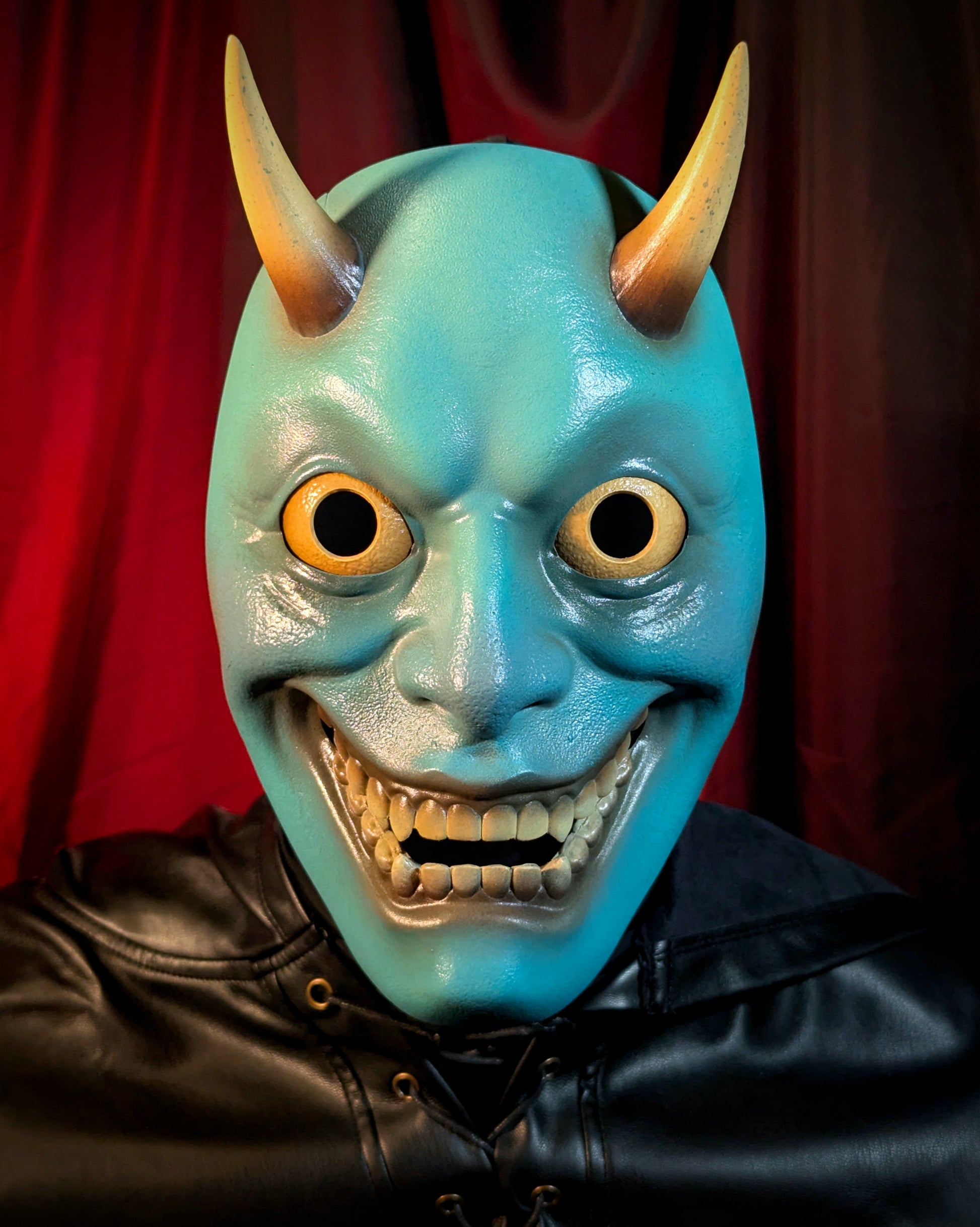 A front facing view of a person wearing a blue hannya mask with yellow eyes and horns against a red curtain background