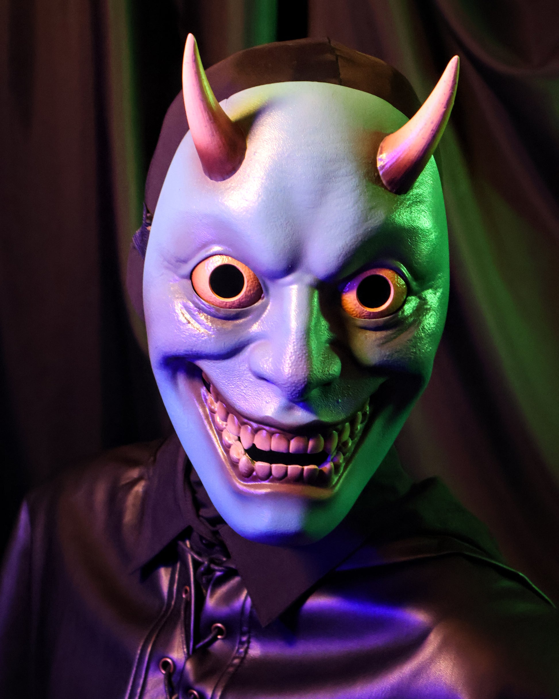 A closeup of a person wearing a blue hannya mask with yellow horns, eyes, and teeth against a green curtain background