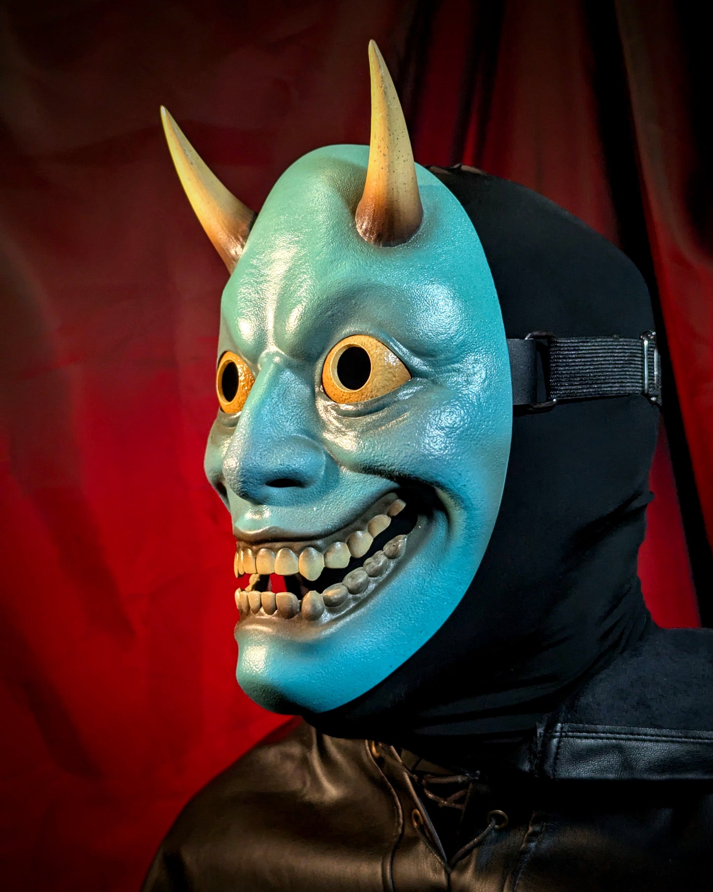 A side angle view of a person wearing a blue hannya mask with yellow horns, eyes, and teeth against a red curtain background