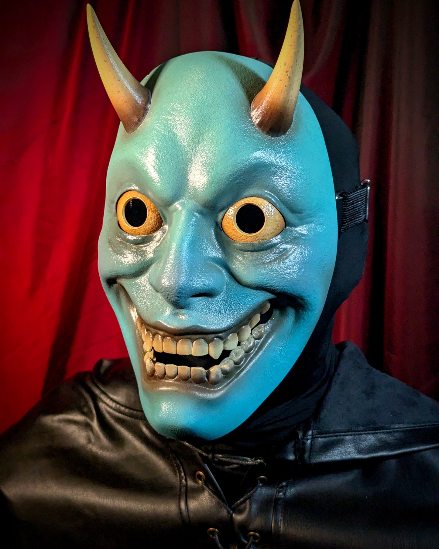 Person wearing a blue hannya mask with large eyes and horns against a red curtain background
