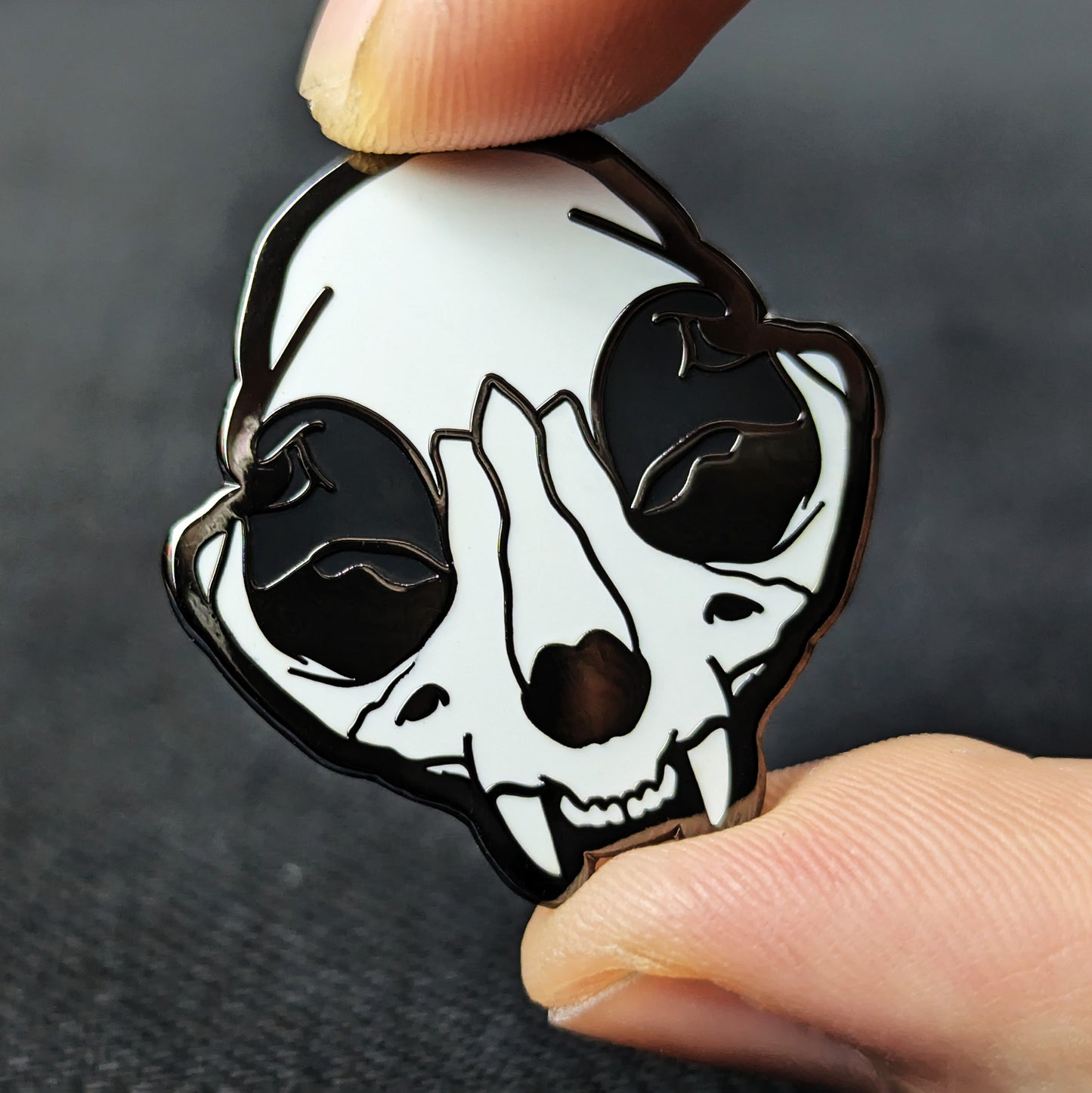 A hard enamel pin with a depiction of a cat skull with black nickel plating, held by a hand.