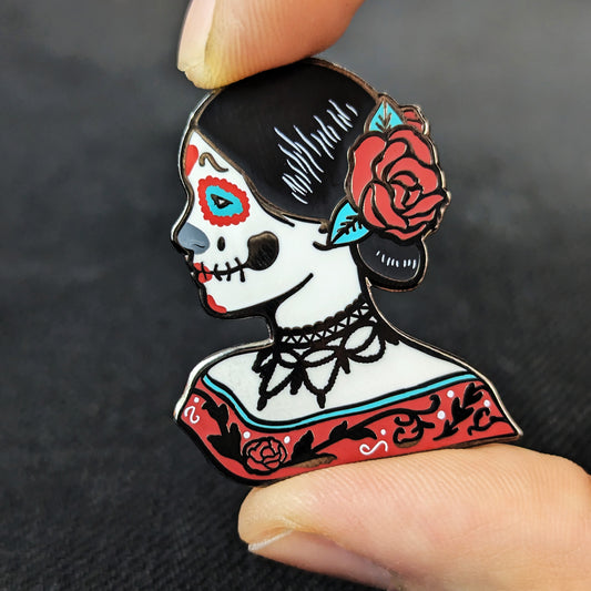 An enamel pin with the design of a woman wearing skeleton makeup with a flower in her hair 
