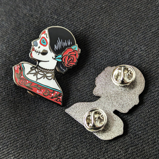 Two enamel pins of a woman with skeleton makeup with one pin facing up and the other facing down on a flat surface