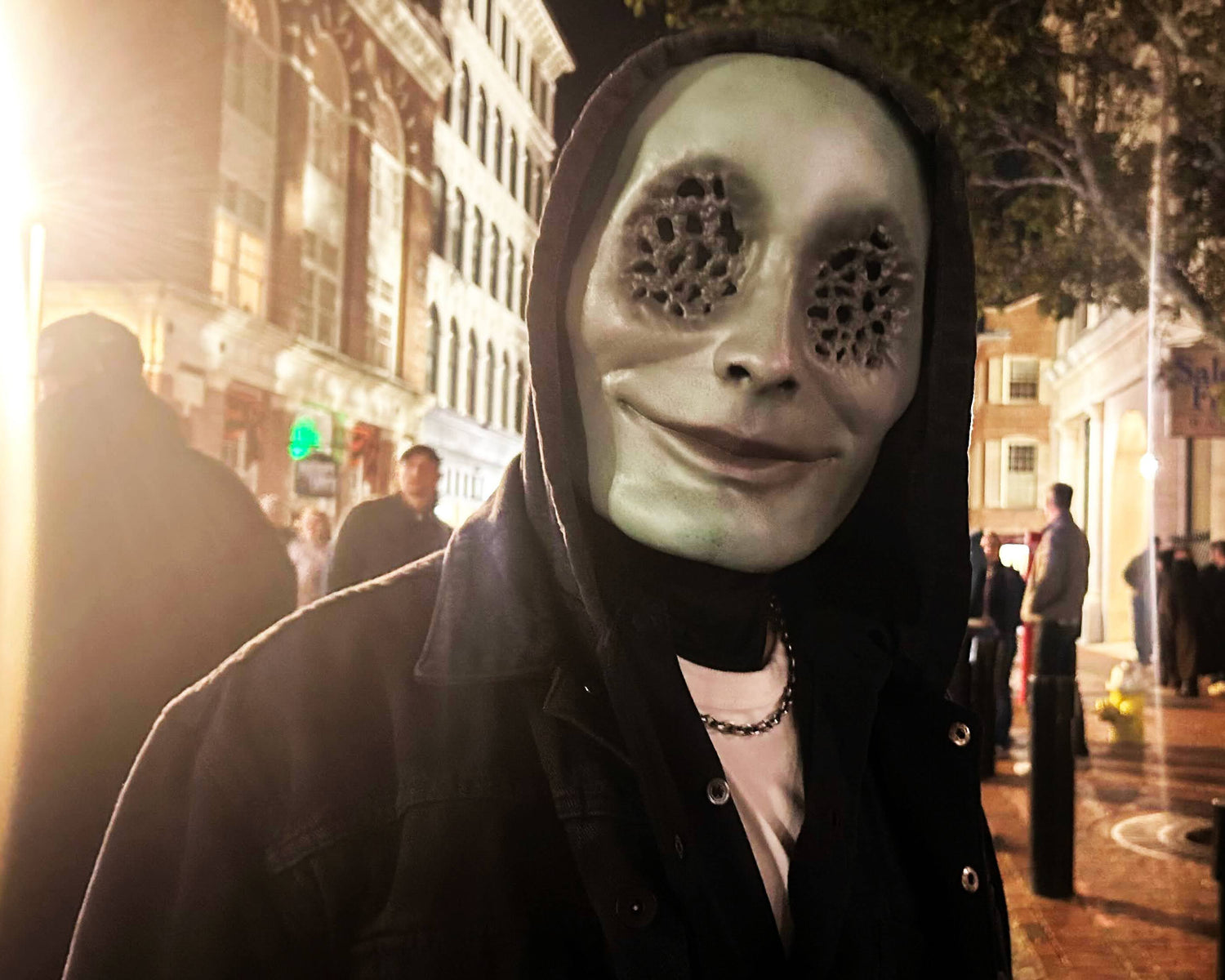 Person wearing a green mask with tiny holes for eyes in a dark hoodie on the street at night