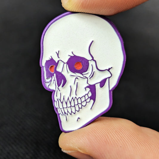 A white soft enamel human skull pin with purple plating and red eyes