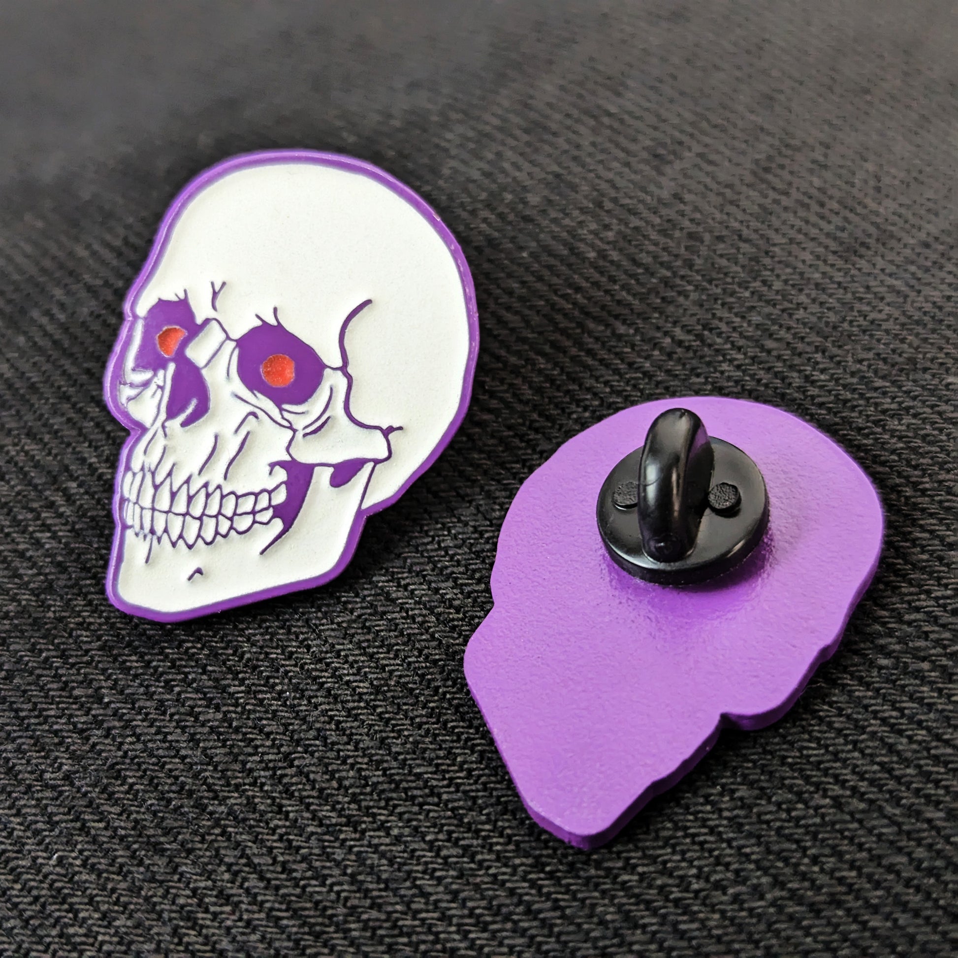 A human skull enamel pin with purple plating and red eyes next to an identical pin facing down to reveal a rubber backing