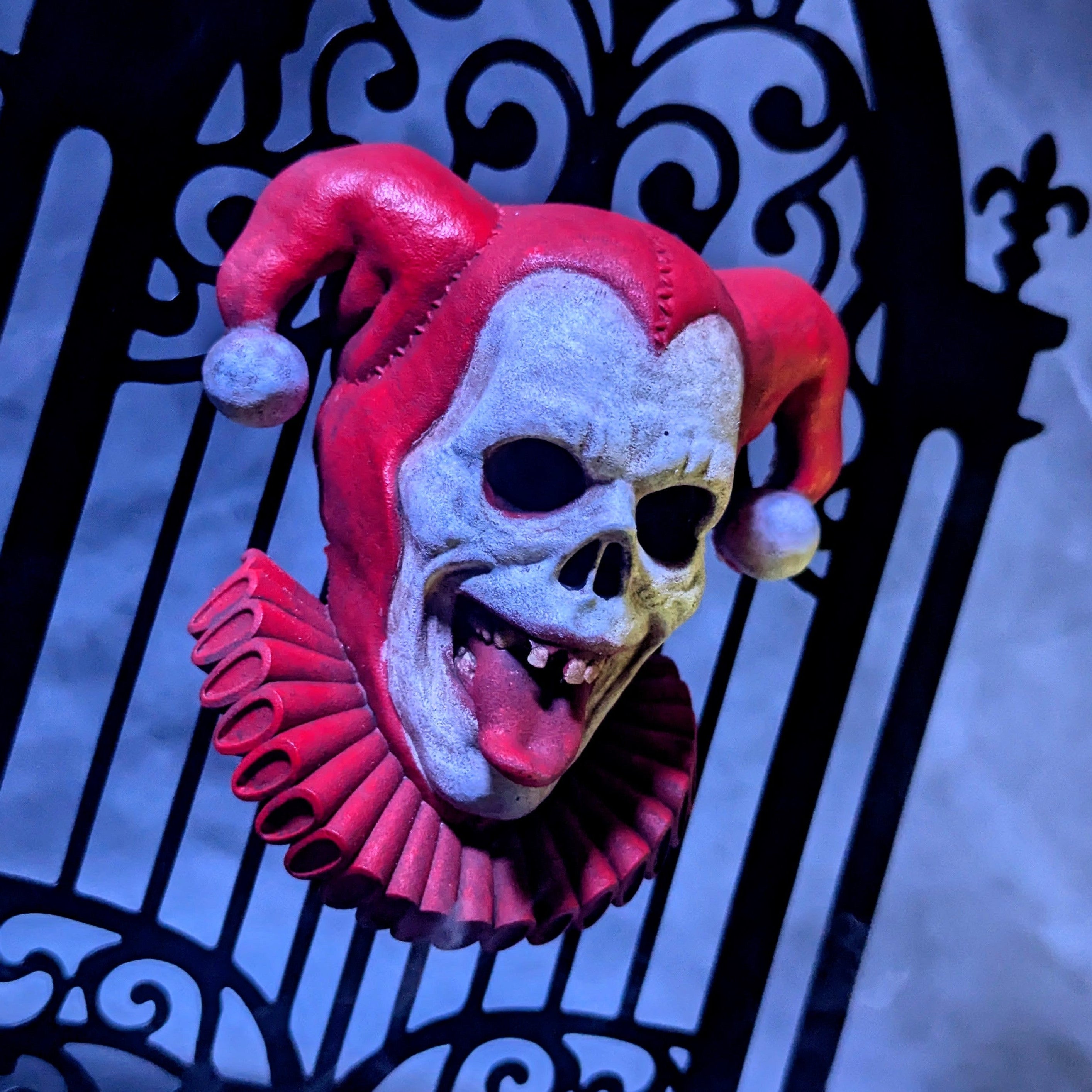 A resin zombie jester magnet with a white face and red costume attached to a small metal gate against a foggy background