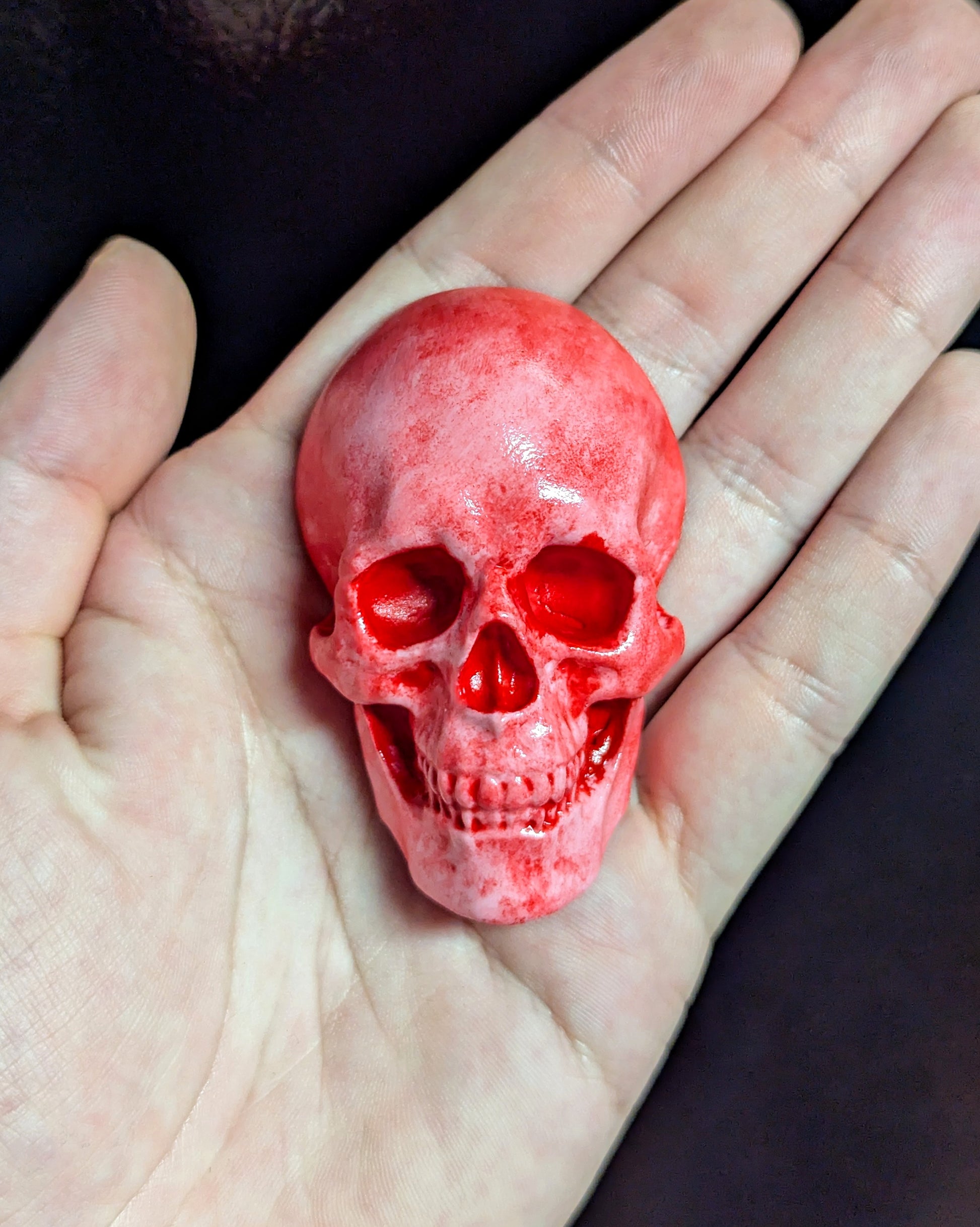 A pink and red miniature skull magnet held in a hand against a dark background