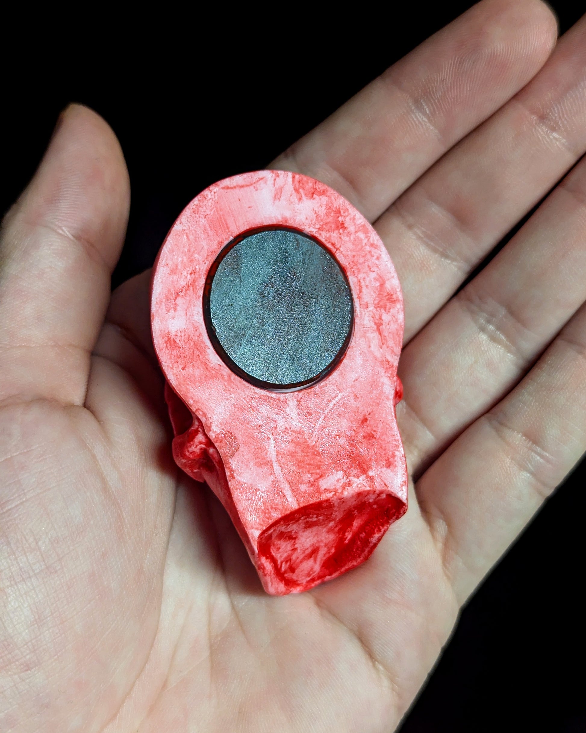 A pink and red miniature skull magnet being held with the magnet side facing outwards against a dark background