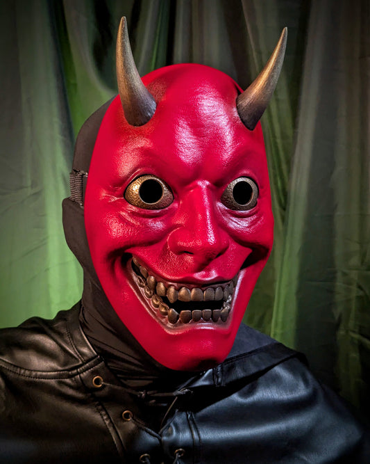 A closeup of a person wearing a red hannya mask with golden horns, eyes, and teeth against a green curtain background