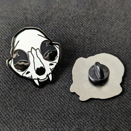 A hard enamel pin with a depiction of a cat skull with black nickel plating next to an identical pin facing downwards showing off a rubber backing
