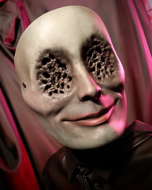 Person wearing a green mask with a faint smile and many small holes for the eyes against a pinkish-red background